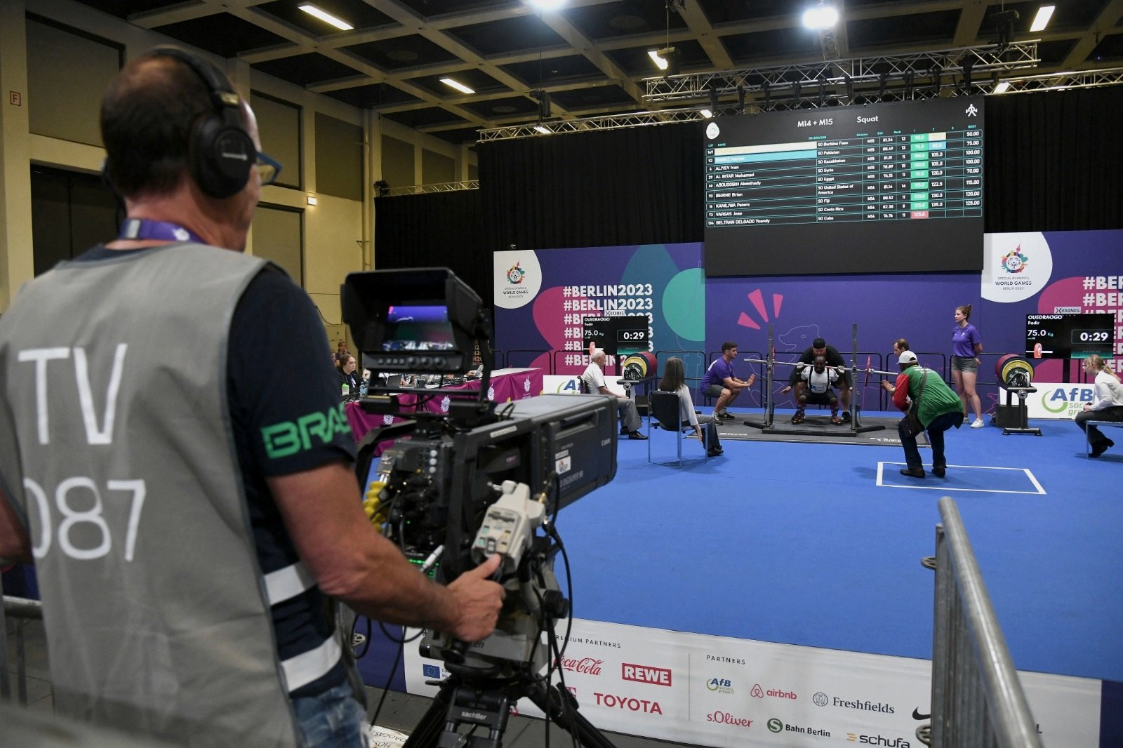 Live competition at the Special Olympics World Games Berlin 2023: a broadcast camera operator in the foreground, the results board above the competition floor