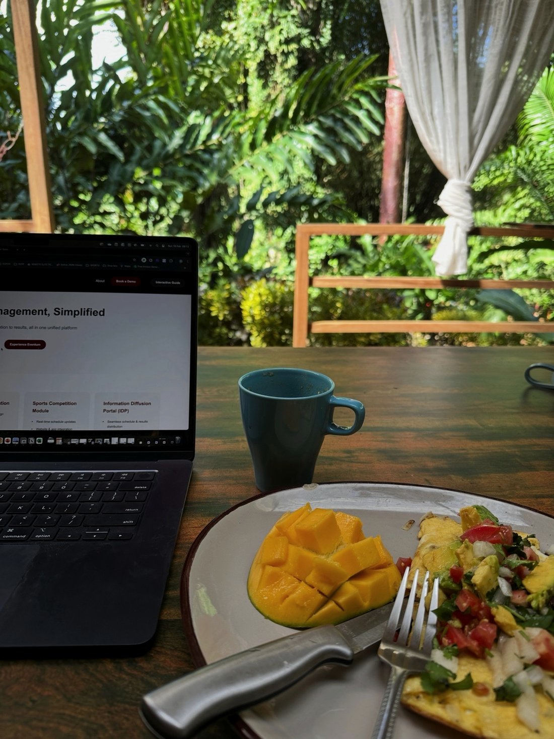 A laptop open beside breakfast, mango and coffee, on a veranda facing tropical garden